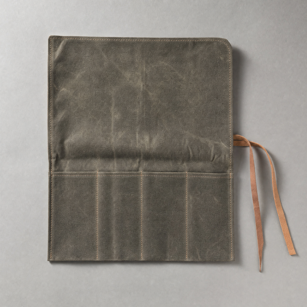 The four-slot knife roll unrolled flat in olive-grey waxed canvas, its four stitched slots empty, with two leather ties loose beside it.
