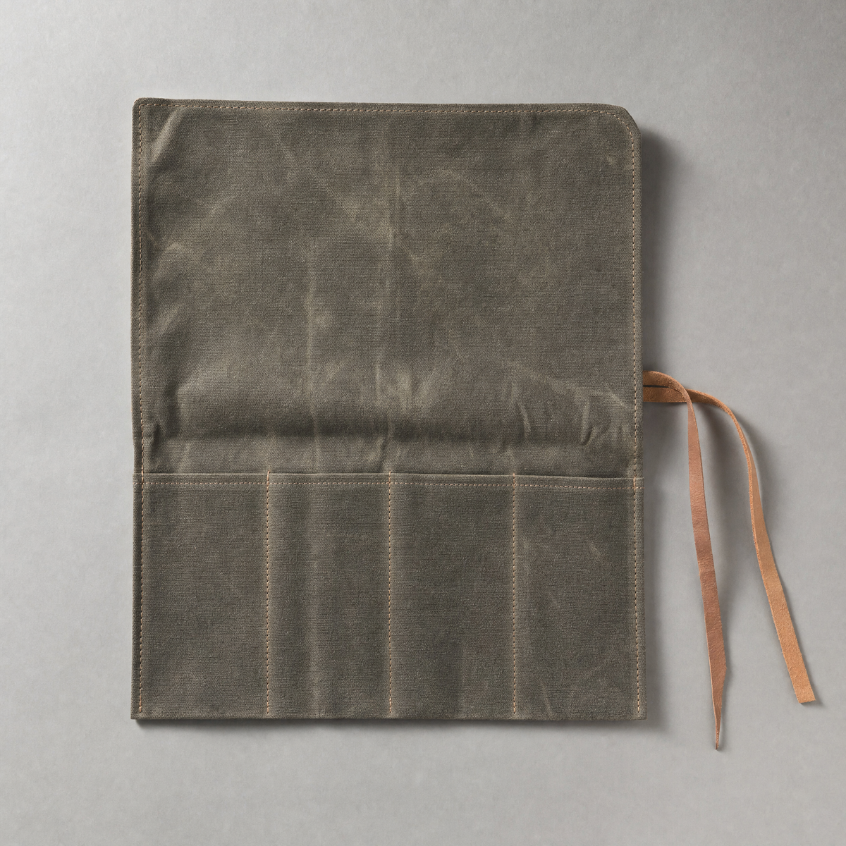 The four-slot knife roll unrolled flat in olive-grey waxed canvas, its four stitched slots empty, with two leather ties loose beside it.